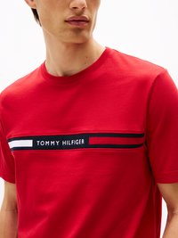 Rotes Baumwoll-T-Shirt mit rundem Halsausschnitt, ausgestattet mit einem horizontalen schwarzen Streifen mit weißen und roten Streifen und dem Text "TOMMY HILFIGER" in Weiß.