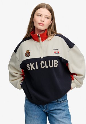 Ung kvinde med langt brunt hår iført en mørkeblå, hvid og rød Ski Club-pullover med halvlukning og blå jeans, står med hænderne i lommerne.