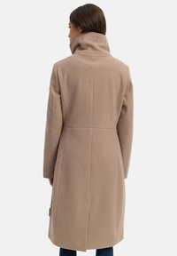 Langer beiger Mantel mit hohem Kragen, ausgestattet mit einer Rückennaht und seitlichen Taschen. Hergestellt aus einem weichen, strukturierten Stoff mit subtiler Markierung.