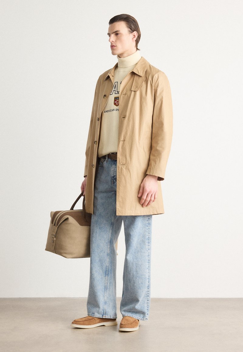 Manteau beige léger sur un pull à col roulé crème, jeans en denim bleus et chaussures marron. Le mannequin tient un sac beige avec des accents marron.