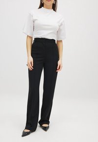 Weißes kurzärmliges Shirt mit hohem Kragen, kombiniert mit schwarzen, hoch geschnittenen, weit geschnittenen Hosen. Schwarze spitze Absatzschuhe runden das Outfit ab.