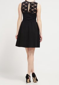 Schwarzes Kleid mit tailliertem Oberteil und ausgestelltem Rock, mit einem Spitzenrücken und einem Schlüsselloch-Ausschnitt. Das Modell trägt schwarze High Heels.