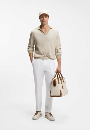 Mann trägt beigen Strickpullover, weiße Hose, weiße Kappe und Turnschuhe, hält eine große beige-braune Handtasche an den Griffen.