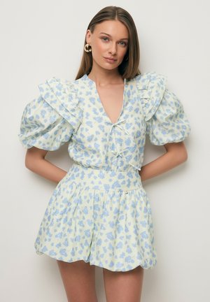 Vrouw draagt een kort bloemenjurkje in lichtgeel en blauw met opgeblazen mouwen en ruches, staand tegen een effen lichte achtergrond.