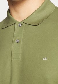 Polo vert olive en tissu texturé, avec col, deux boutons et petit logo "ck" blanc brodé à l'ourlet.