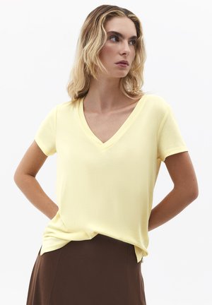 Kobieta z falującymi blond włosami, ubrana w jasnof żółty top z krótkim rękawem i dekoltem w serek, włożony do brązowej spódnicy, patrząca w lewo z rękami za plecami.