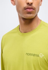 Bărbat purtând un tricou cu mâneci lungi Norrøna de culoare verde lime, vizibil de la gât până la piept, cu accent pe logo-ul brandului pe partea stângă.