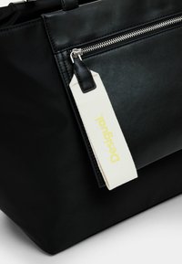 Black leather tote bag featuring a silver zipper and a white tag with "Desigual" printed in yellow. Smooth texture with minimalist design.