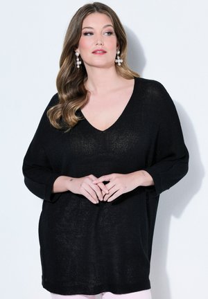 Kvinde med langt, bølget hår iført perleøreringe med dråbeform og en løs sort V-halstrøje, stående foran en hvid baggrund.