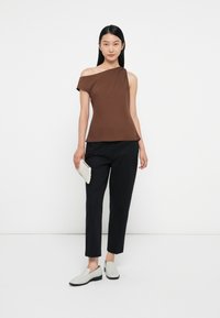Barna aszimmetrikus felső egy vállra lecsúszó ujjal, fekete szűkített nadrággal és világosszürke slip-on cipővel párosítva. Texturált fehér clutch-t tart a kezében.
