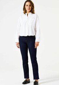 Chemise blanche à col boutonné avec un ourlet arrondi ; jeans droits bleu foncé ; mocassins foncés ; modèle se tenant contre un fond neutre.
