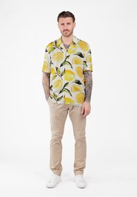 Hombre con tatuajes que lleva una camisa de manga corta con estampado de limones amarillos, pantalones beiges y zapatillas blancas, de pie con las manos en los bolsillos.