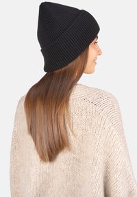 Bonnet noir en maille côtelée avec un sommet pointu, porté avec un pull crème en grosse maille, laissant apparaître de longs cheveux bruns tombant dans le dos.
