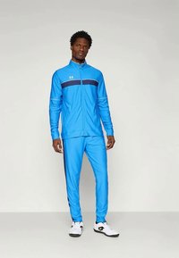 Άνδρας που στέκεται φορώντας φωτεινό μπλε στήθος με σκούρα ρίγα στην περιοχή του στήθους και λευκά αθλητικά παπούτσια σε απλό φόντο.