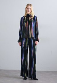Donna in piedi indossando una blusa nera in satin morbido e pantaloni larghi a righe verticali multicolori, con scarpe col tacco nere.