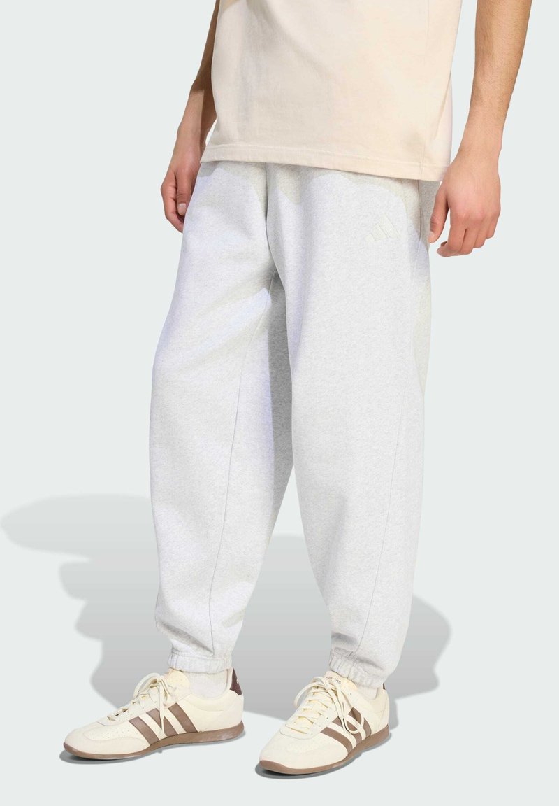 Personne portant un pantalon de survêtement Adidas gris clair et des baskets beiges avec des bandes marron, debout sur un fond gris clair.