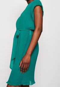 Turkos veckad klänning med korta ärmar, ett midjeband och en scalloped nederkant. Tillverkad av lättviktig tyg, designad för en avslappnad passform.