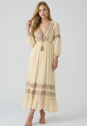 Joven mujer con un vestido largo de manga larga color crema con bordados marrones y cordón con borlas, de pie frente a un fondo claro y liso.
