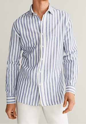Homme portant une chemise à manches longues à rayures verticales bleues et blanches avec un pantalon blanc sur un fond neutre.