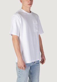 T-shirt bianca in cotone con scollatura rotonda, maniche corte e taschino sul lato sinistro del petto. Tessuto morbido, vestibilità leggermente ampia, abbinata a jeans azzurri chiari.