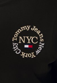 Czarna tkanina z okrągłym haftowanym logo z napisem "Tommy Jeans", "NYC" i "New York City", w towarzystwie małej flagi w trzech kolorach.