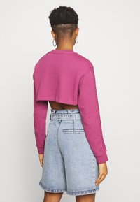 Person med kort hår och hoopörhängen, iklädd en kortärmad rosa topp och högmidjade ljusa denimshorts, sedd bakifrån.