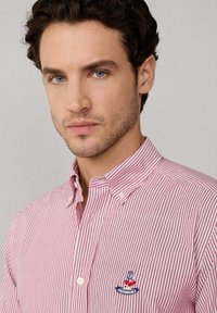 Camisa de botones a rayas rojas y blancas con cuello clásico, bolsillo frontal con un logotipo bordado y tejido texturizado.
