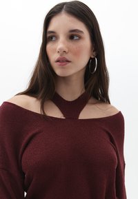 Weinroter Strickpullover mit schulterfreiem Design und einem Schlüsselaugen-Detail am Hals, ausgestattet mit einem strukturierten Stoff und langen Ärmeln.