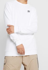 Sudadera blanca de manga larga con puños acanalados. Presenta un logo negro de Adidas en el pecho. Fabricada en una suave tela texturizada.