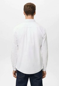 Camisa blanca de manga larga con un corte relajado, cuello clásico, canesú trasero y puños abotonados, combinada con vaqueros de mezclilla oscura.