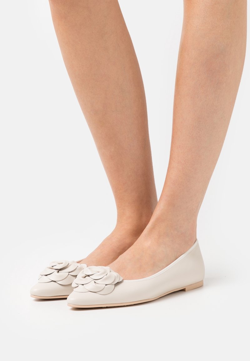 Pieds portant des ballerines blanches en cuir ornées de fleurs superposées sur les bouts pointus, sur un fond blanc uni.