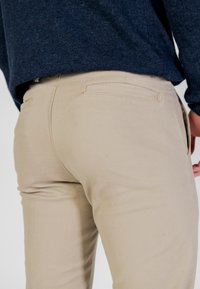 Pantalones de algodón beige con bolsillos traseros, mostrados junto a un suéter azul marino. La tela es suave con un ligero brillo, de corte ajustado.