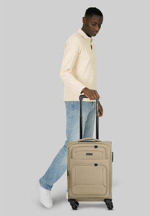 Junger Mann in beigem Pullover und Jeans zieht einen beigen Koffer mit Teleskopgriff vor grauem Hintergrund.