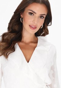 Fehér, virágmintás mintázatú átlapolt blúz fodros nyakkivágással és hosszú ujjakkal; finom aranylánc nyaklánccal és karikaalakú fülbevalóval párosítva.