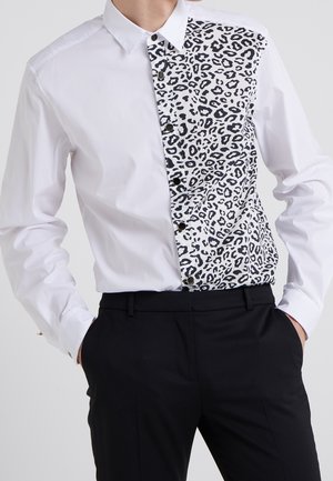 Hvid langærmet skjorte med sort-hvid leopardprintpanel på den ene side, kombineret med sorte bukser og hænderne i lommerne.