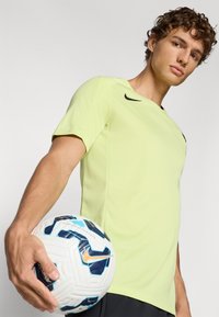 Camisa atlética Nike de color verde claro con mangas cortas, que presenta un logo negro. El modelo sostiene un balón de fútbol blanco con acentos azules y naranjas.