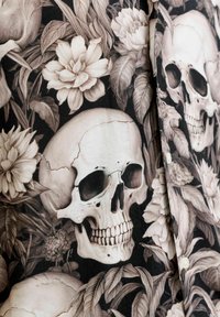 Crânes humains blancs entourés de grandes fleurs blanches et de feuilles sombres sur un tissu foncé, avec des boutons visibles le long du bord.