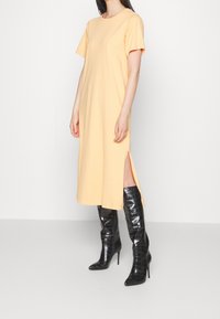 Żółta sukienka z bawełny w kształcie T-shirtu z krótkimi rękawami i bocznymi rozcięciami, połączona z błyszczącymi, czarnymi, wysokimi na kolana botkami na szpilce. Prosty design, casualowy krój.
