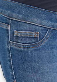 Un jean en denim bleu présente une petite poche arrière avec des coutures orange, et une texture lisse avec une finition légèrement décolorée près de la taille.