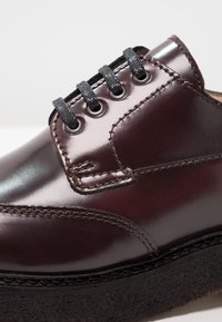 Chaussure en cuir bordeaux avec une finition brillante, bout rond et lacets noirs. Présente des coutures visibles et une semelle noire texturée.