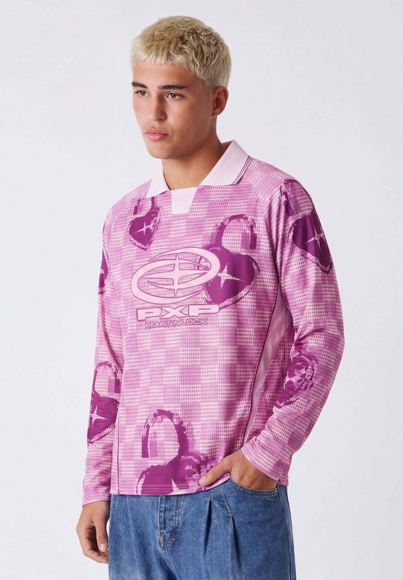 Jeune homme aux cheveux blonds courts portant une chemise rose à manches longues avec des motifs de cœurs et de cadenas, ainsi qu'un jean bleu, debout devant un fond blanc.