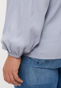 Blouse bleu clair à manches longues avec des rayures verticales, des poignets froncés et une texture lisse, associée à un jean bleu. Une main visible repose sur la hanche.