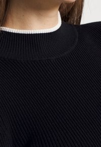 Zwarte geribde trui met een hoge nek en een contrasterende witte band. De textuur is zacht en gestructureerd, met verticale details.