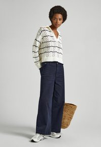 Adīts džemperis ar atvērto rakstu krēmkrāsā, ar tumši zilu svītru; kopā ar platām kājām tumši zilās bikses un baltām kedām. Pusē ir austs soma.