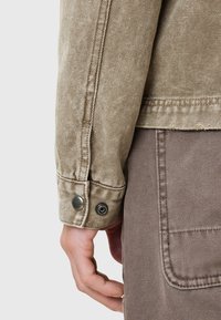 Gros plan sur le bras d'une personne portant une veste beige texturée avec des boutons-pression métalliques et un pantalon marron avec une grande poche arrière.