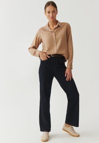 Beige satijn shirt met knopen, gecombineerd met donkerblauwe wijde jeans en lichtbeige sneakers. Eenvoudig ontwerp, klassieke pasvorm en gladde textuur.