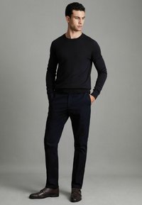 Un pull en maille bleu marine et un pantalon bleu marine ajusté complètent la tenue, associés à des chaussures en cuir marron foncé à double boucles.