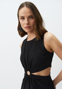 Robe noire sans manches avec un détail de nœud à la taille et des ouvertures sur les côtés, dotée d'un accent rond. Fabriquée en tissu texturé.