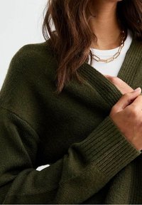 Cardigan lavorato a maglia verde oliva con polsini a coste e apertura frontale, indossato sopra una maglietta bianca, con texture liscia. Collana in oro visibile.