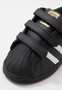 Mustat lenkkarit, joissa on kolme tarranauhaa, kuminen kärki, valkoiset raidat ja kultainen logo sivussa. Pintarakenteeltaan ja sileästä nahasta valmistettu materiaali.